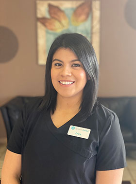 This is a photograph of a woman standing indoors, wearing a short-sleeved dark top and a name tag with a light background. She has short hair and appears to be smiling at the camera. The setting suggests she may be in a professional environment, possibly an office or waiting area.