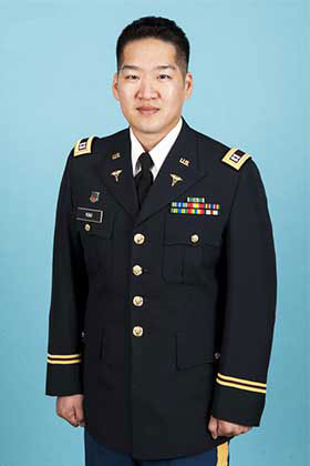 The image shows a man in military attire, standing against a blue background.