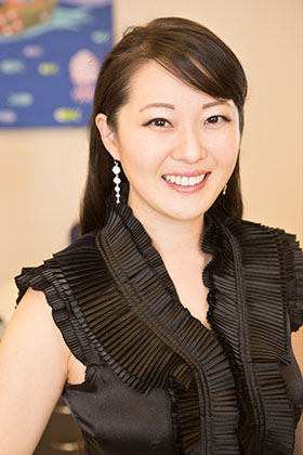 A woman wearing a white lab coat and mask is smiling at the camera, seated in a dental office setting.