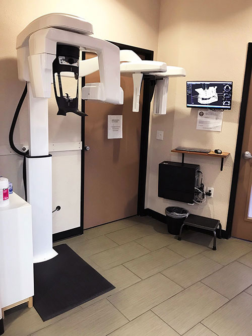 The image shows a dental office with a modern dental chair and equipment, including a digital display screen, situated in front of a large mirror.