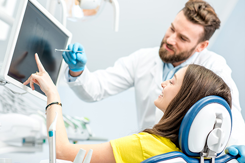 A smiling woman is seated in a dental chair, while a dentist in a white coat stands behind her, holding a tablet with a digital image displayed, likely explaining treatment options.